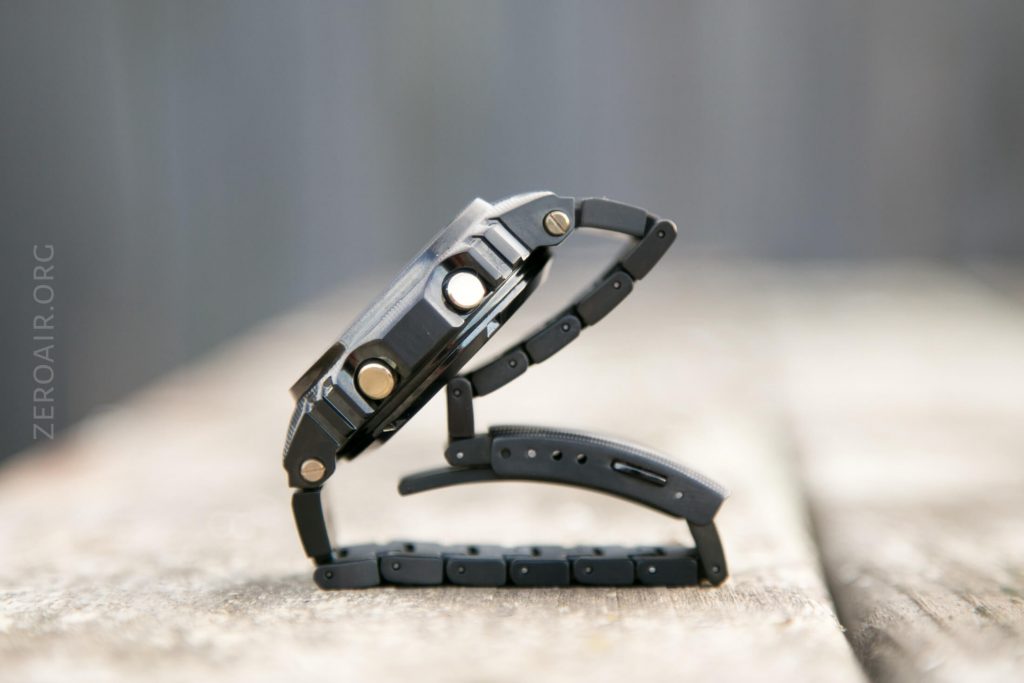 A black digital wristwatch is propped on its side on a wooden surface, with the band partially unfastened and curved so that the watch forms a shape resembling the letter C. The background is blurred.