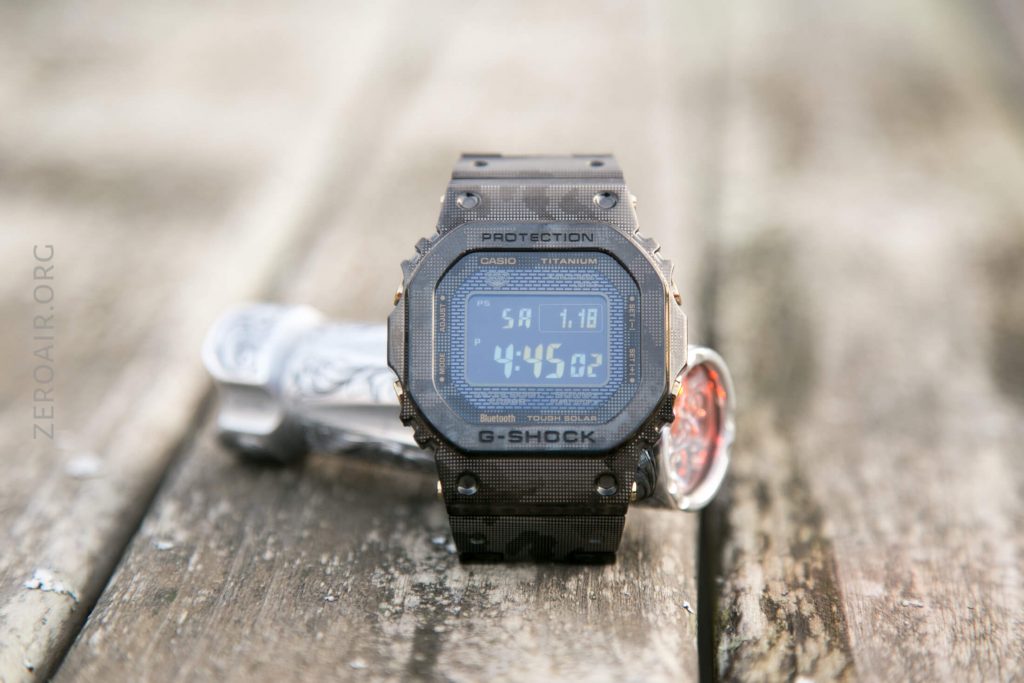 A Casio G-Shock titanium digital watch with a camouflage pattern is displayed upright on a weathered wooden surface, with the screen showing the time 4:45:02 and the date 7/18.