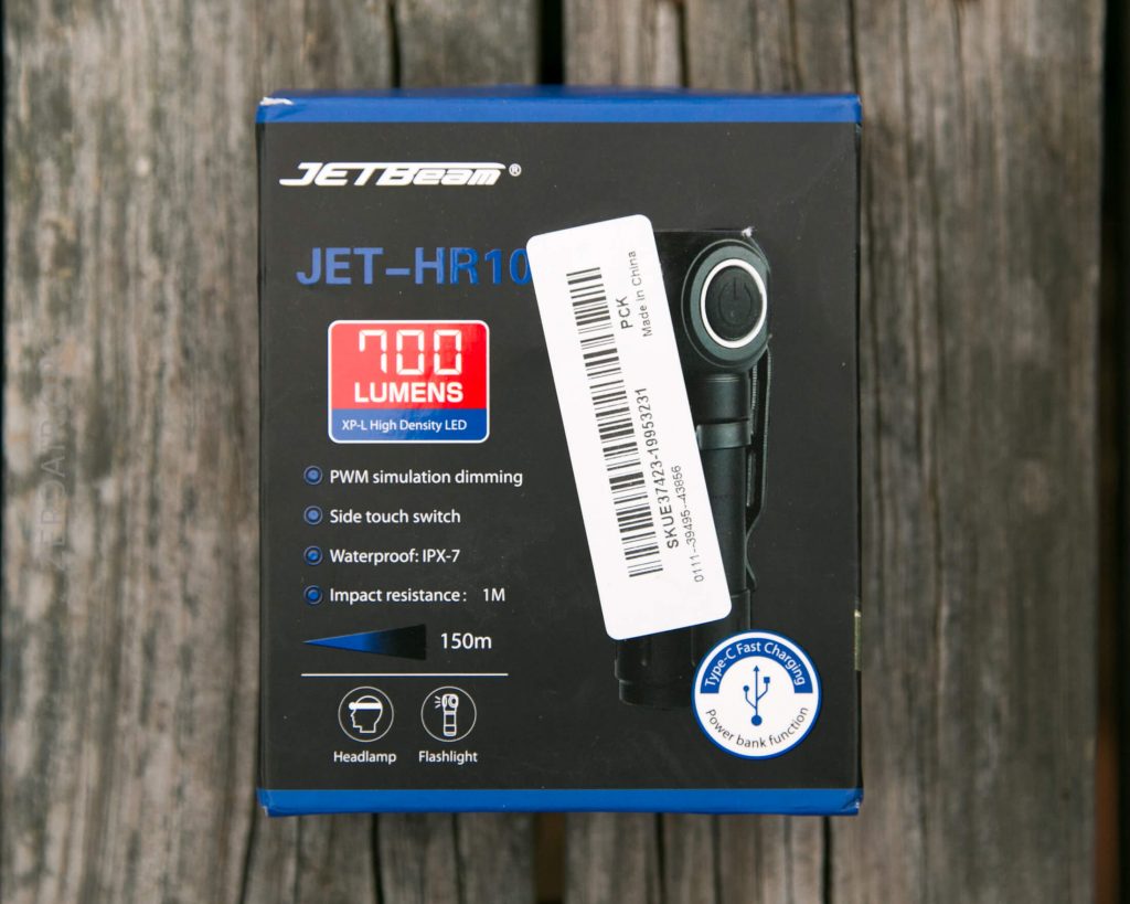A JetBeam JET-HR10 flashlight box is shown on a wood surface. The box lists features like 700 lumens, waterproof IPX-7 rating, 150m impact resistance, and includes a barcode sticker partially covering the text.