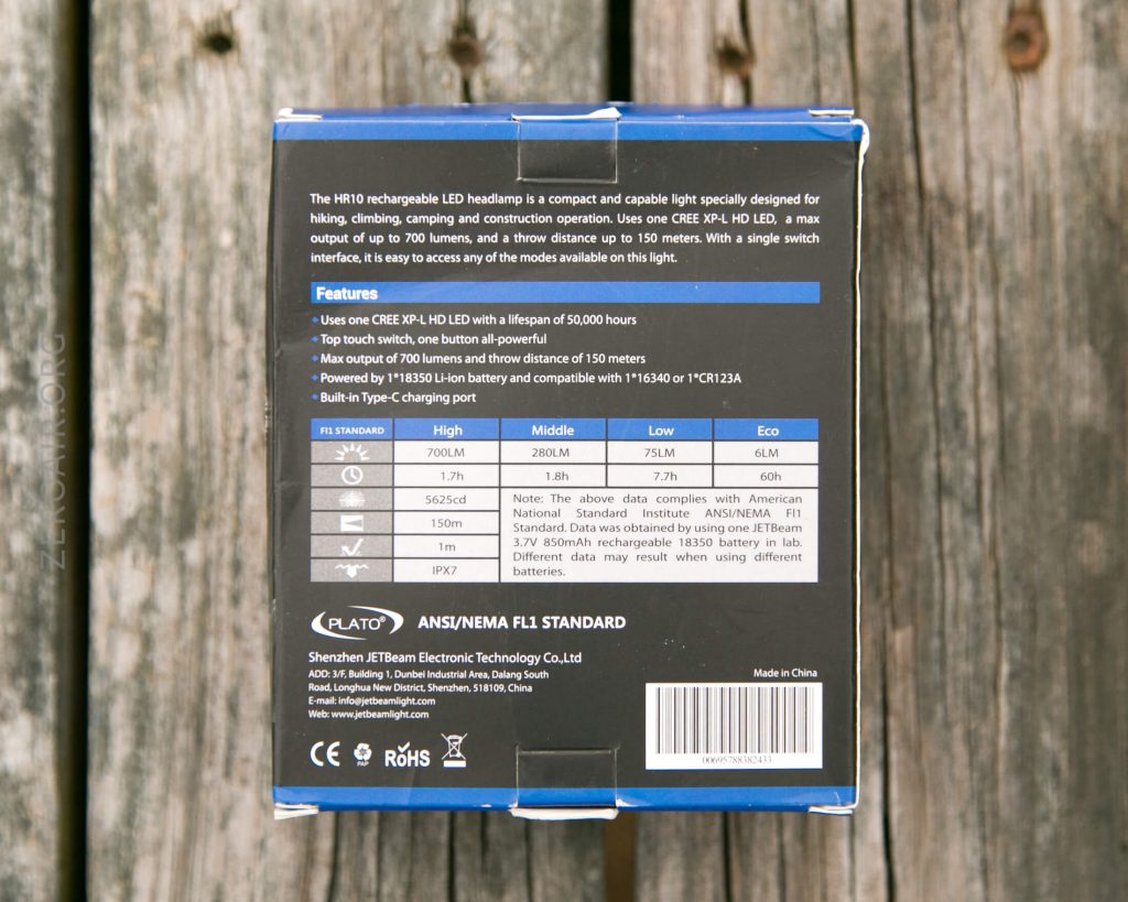 A product box for a rechargeable LED headlamp sits on a wooden surface. The back of the box lists features, technical specs, performance data, and manufacturer details in English.
