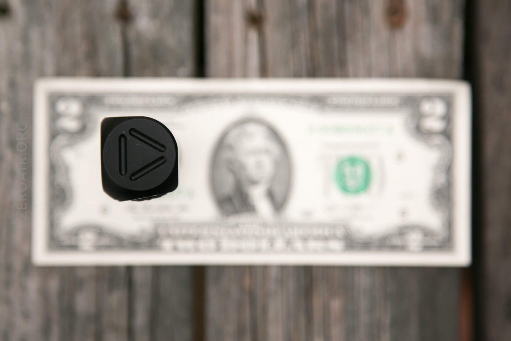 A two-dollar bill lies on a wooden surface, with a black object featuring engraved geometric shapes positioned above it, partially obscuring the bill’s center. The image is photographed from above.