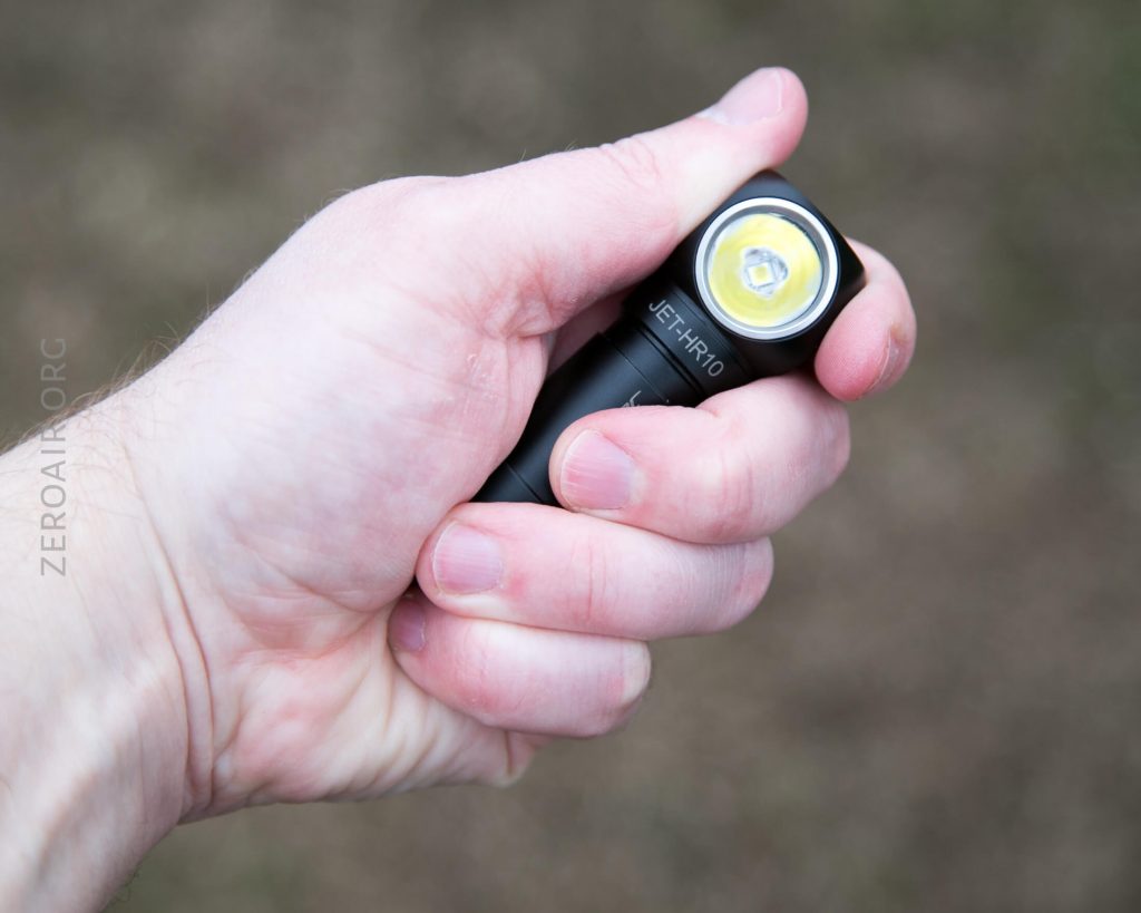A close-up of a hand holding a small black flashlight with a round yellow and white LED, outdoors against a blurred background. The flashlight is labeled “JET HR10” on the side.