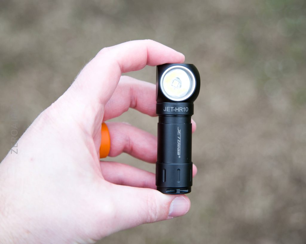 A person holds a compact black flashlight labeled JET-HR10 between their thumb and fingers against a blurred brown background.