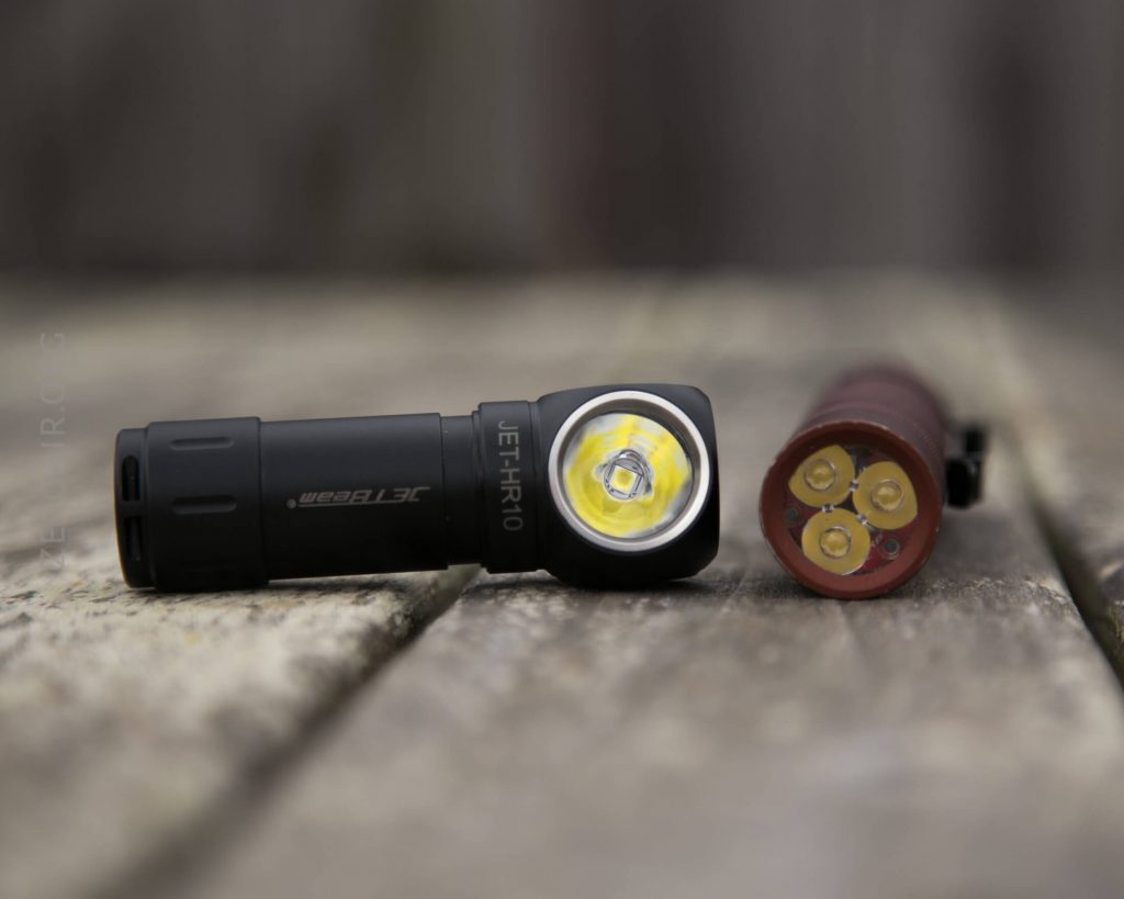 A black right-angle flashlight and a cylindrical three-LED battery pack are lying on a weathered wooden surface, shown in close-up with a shallow depth of field.