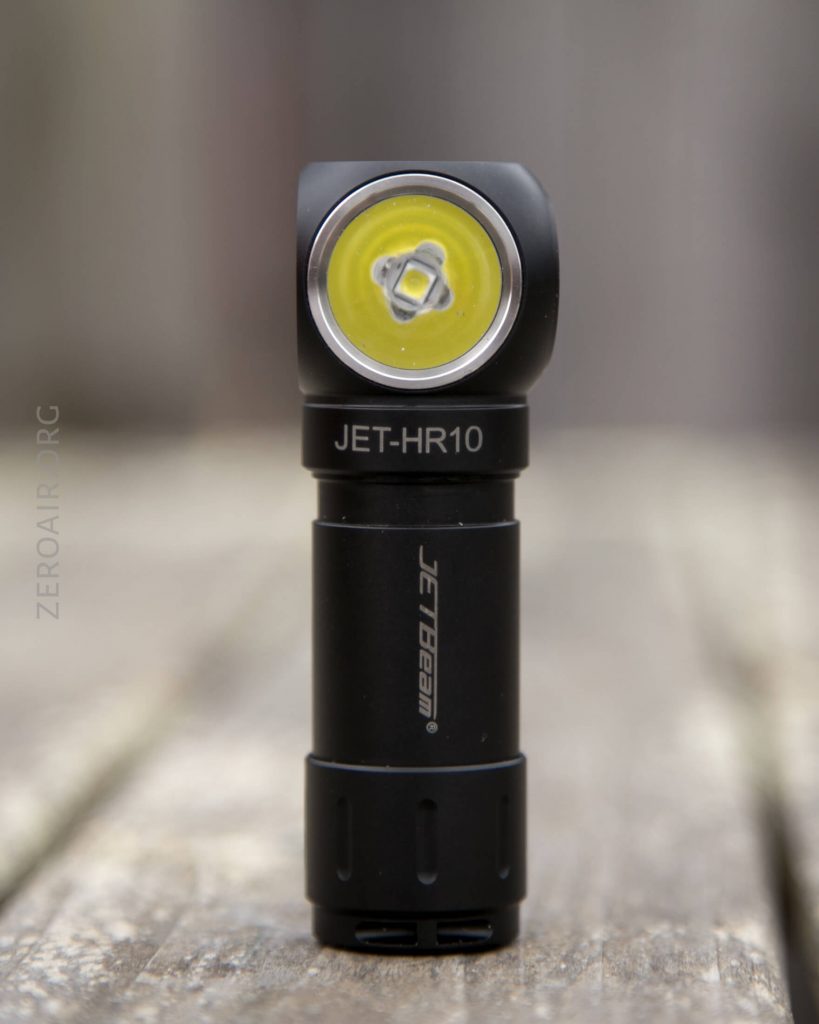 A black JETBeam JET-HR10 angled flashlight stands upright on a wooden surface, with the lens and yellow LED visible facing forward. The background is blurred.