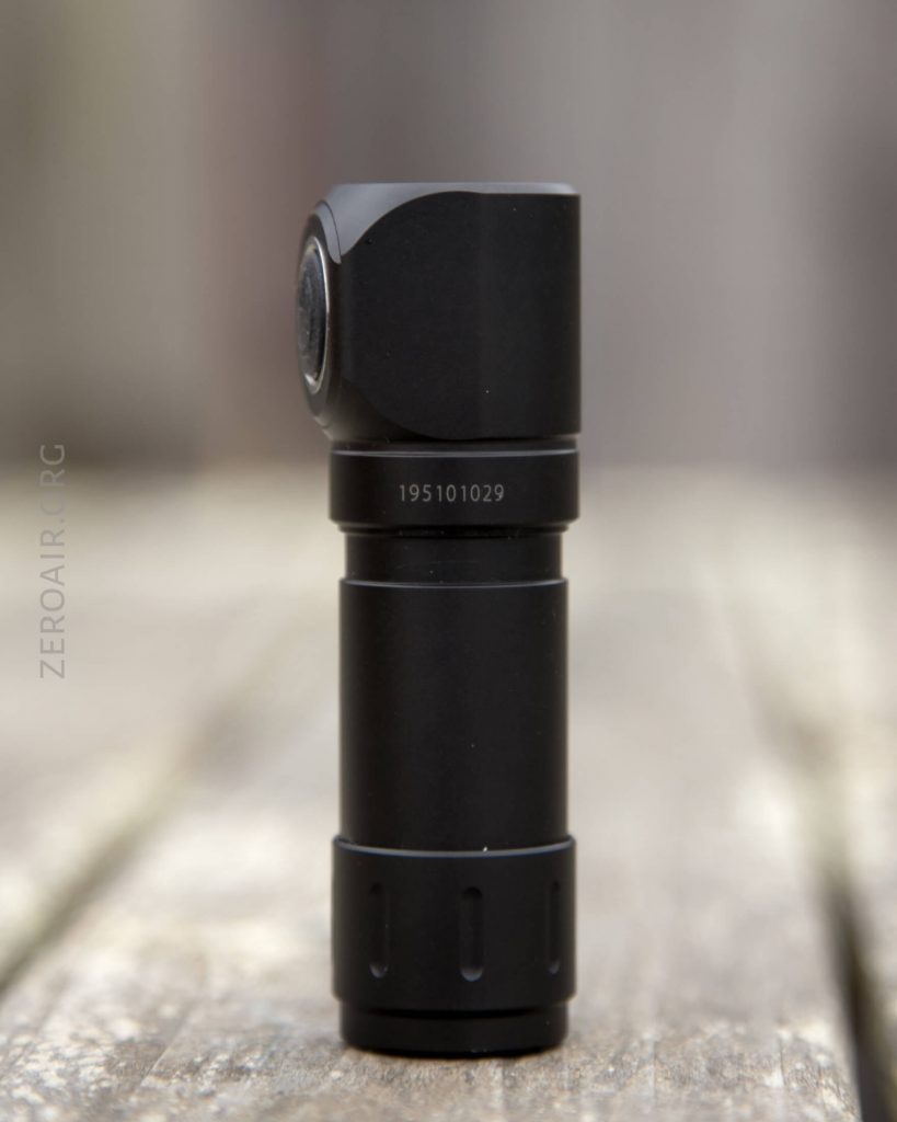A black, cylindrical flashlight with a right-angle head stands upright on a wooden surface. The flashlight has the number 195101029 printed on its side. The background is blurred.
