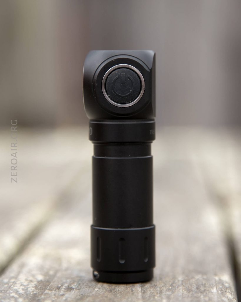 A black, cylindrical flashlight with a right-angle head is standing upright on a wooden surface. The power button is visible on the head of the flashlight. The background is out of focus.