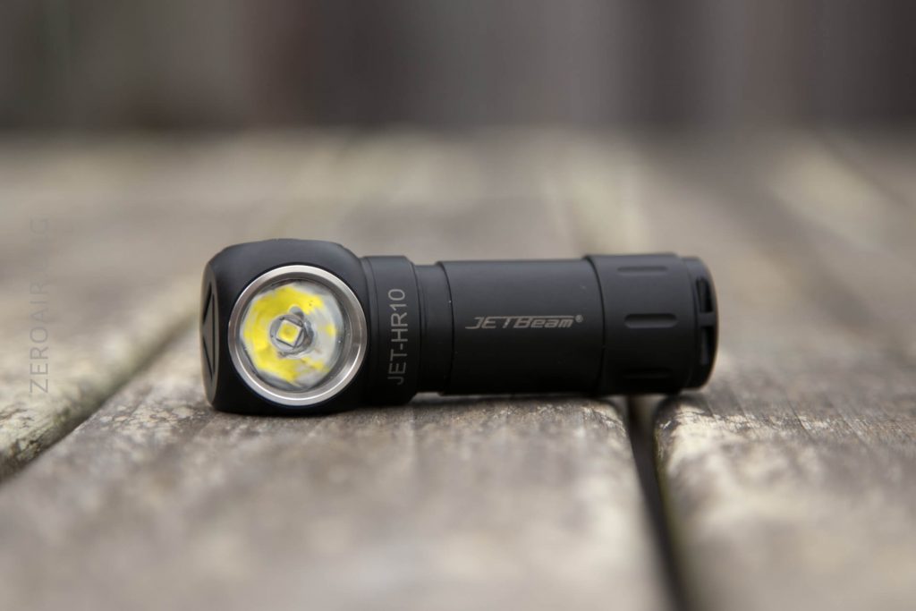 A black JETBeam JET-HR10 flashlight lies on its side on a weathered wooden surface, positioned with the lens facing forward. The background is blurred.