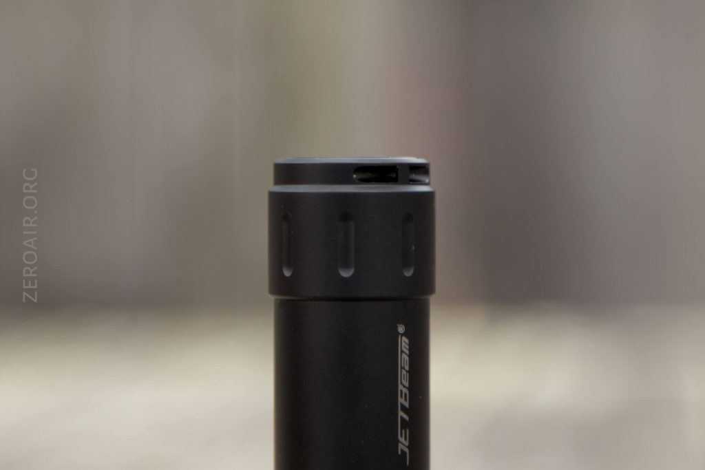 A close-up of the top section of a black cylindrical flashlight with grooves around the top edge. The brand name JETBeam is visible on the side. The background is out of focus.