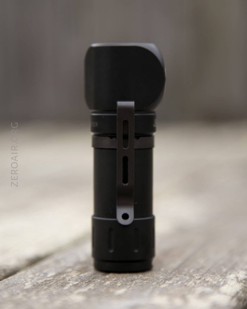 A small black flashlight with a metal clip is standing upright on a wooden surface. The background is blurred, and the text ZEROAIR.ORG is faintly visible along the left side.