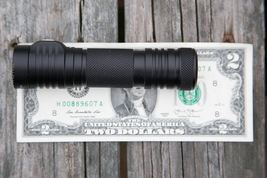 A black flashlight is placed horizontally on top of a U.S. two-dollar bill, which is lying flat on a weathered wooden surface.