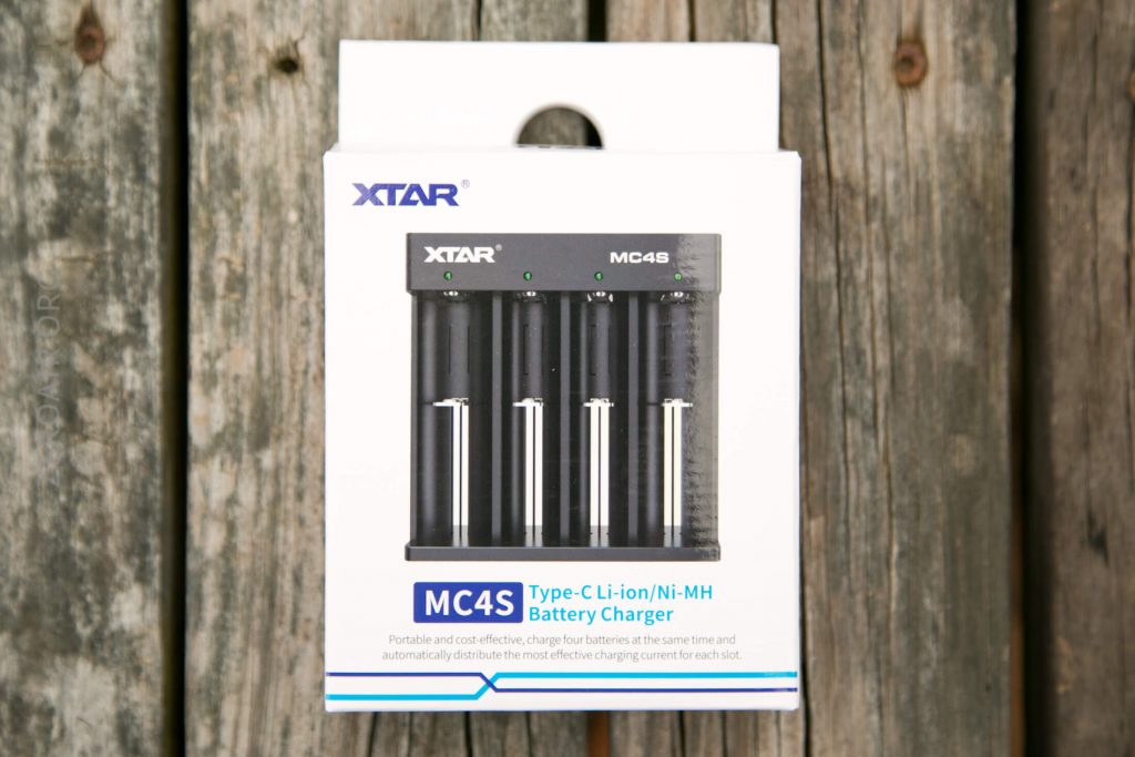 A boxed XTAR MC4S battery charger is shown on a wooden surface. The packaging displays an image of the charger and describes it as a Type-C Li-ion/Ni-MH battery charger for four batteries.