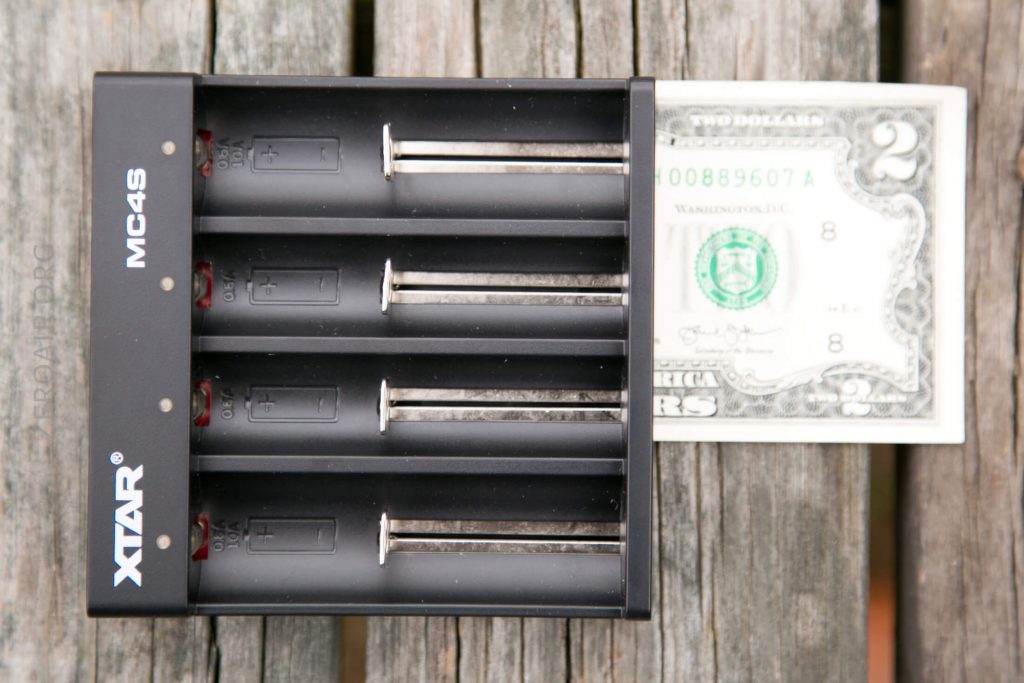 A black XTAR MC4S battery charger with four slots is placed on a wooden surface next to a two-dollar bill for size comparison.