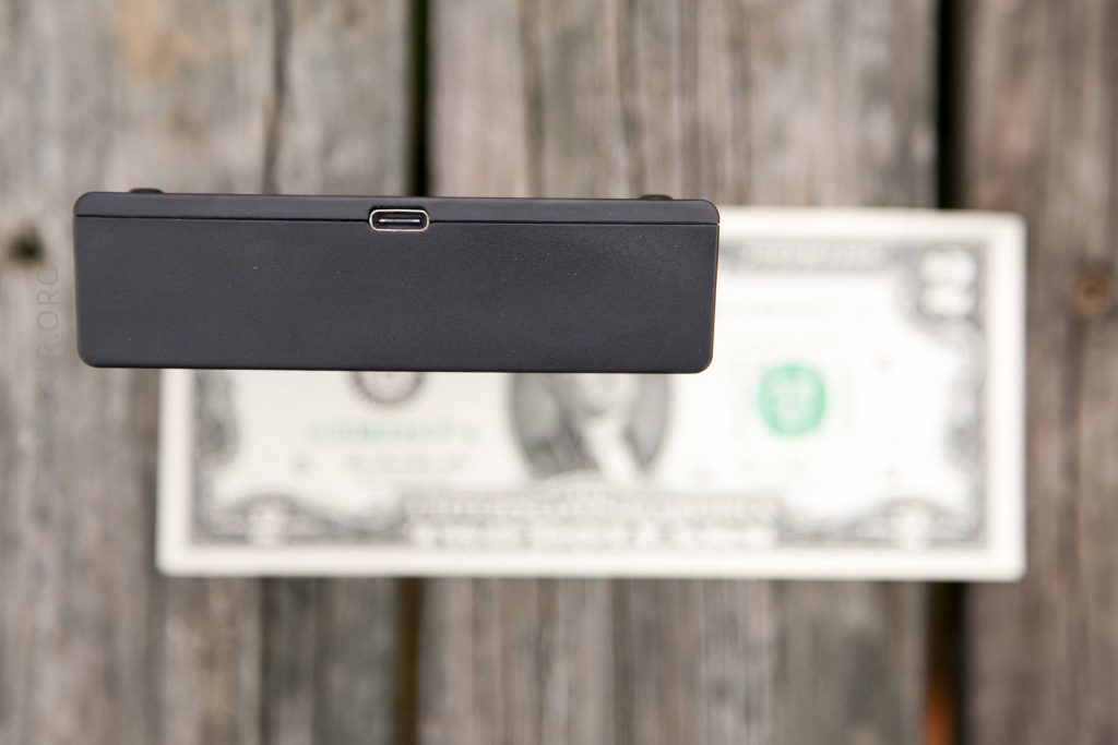 A black rectangular electronic device with a USB-C port is held above a U.S. one dollar bill on a weathered wooden surface.