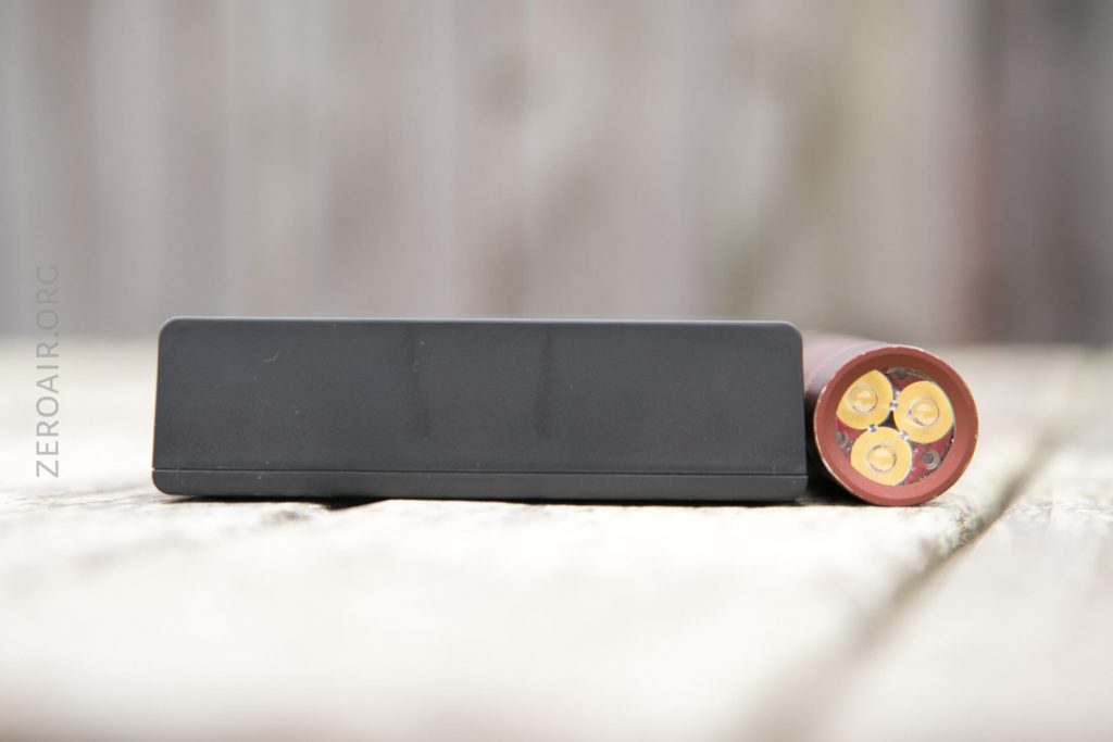 A black rectangular object and a cylindrical battery with three yellow circular contacts on one end are placed side by side on a wooden surface. The background is blurred.