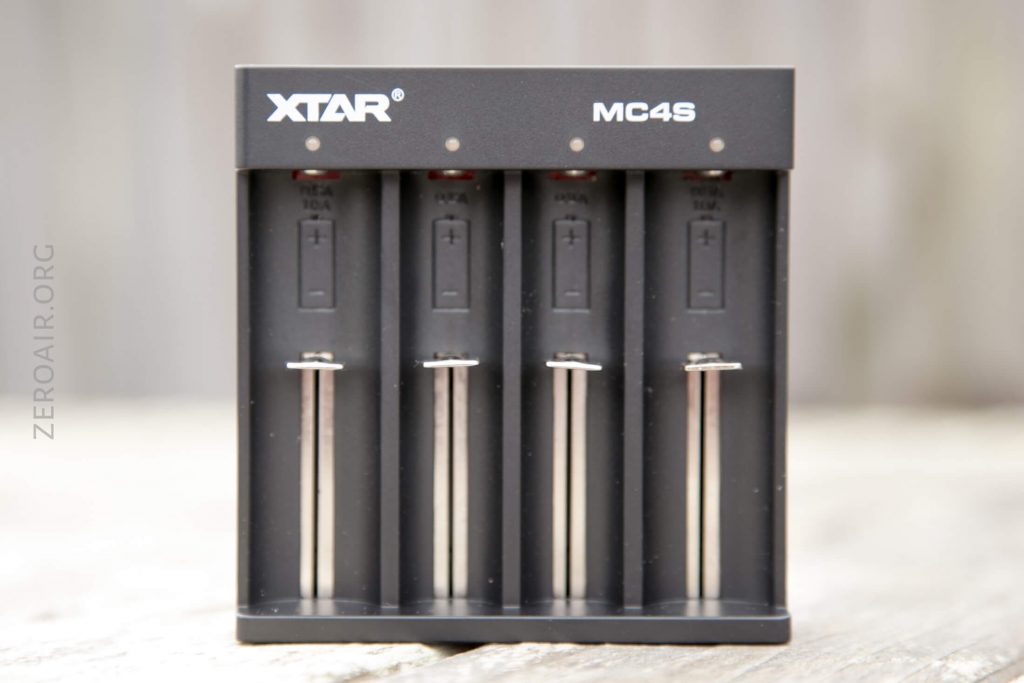 A black XTAR MC4S battery charger with four charging slots, each featuring a spring-loaded contact and labeled indicators for battery polarity, sits on a wooden surface.