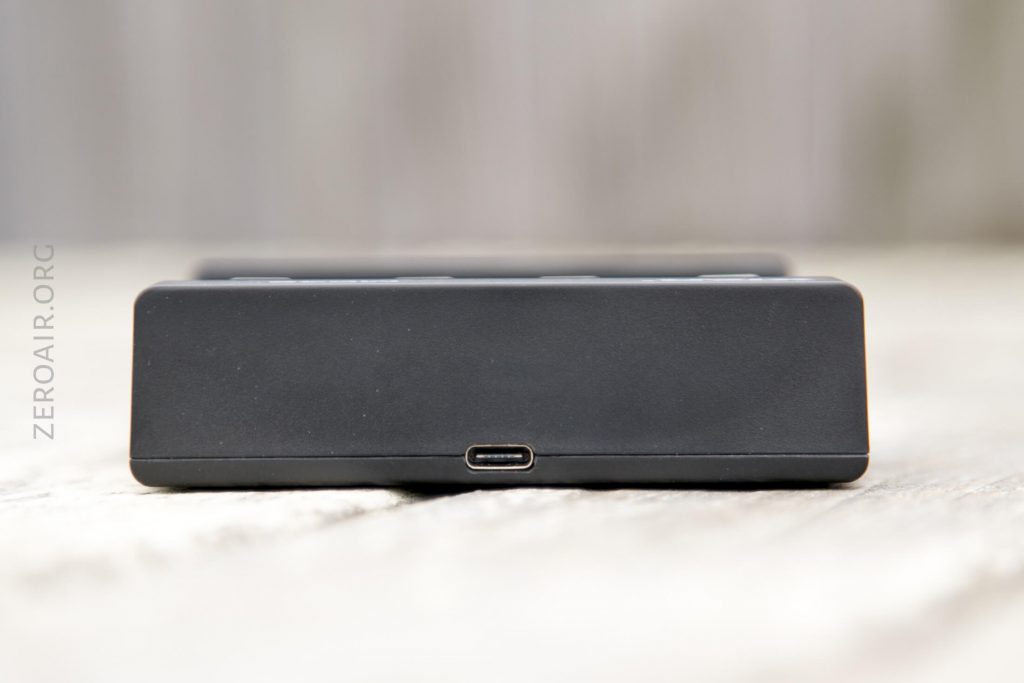 A close-up view of a black rectangular electronic device, showing a single USB-C port in the center of the front side. The background is blurred and the surface appears textured. ZEROAIR.ORG is visible on the left side.
