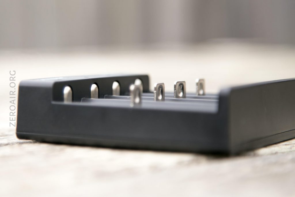 A close-up view of a black battery charger with four metal charging slots. The charger is placed on a light-colored surface, and the background is blurred. The text ZEROAIR.ORG is visible on the left side of the image.