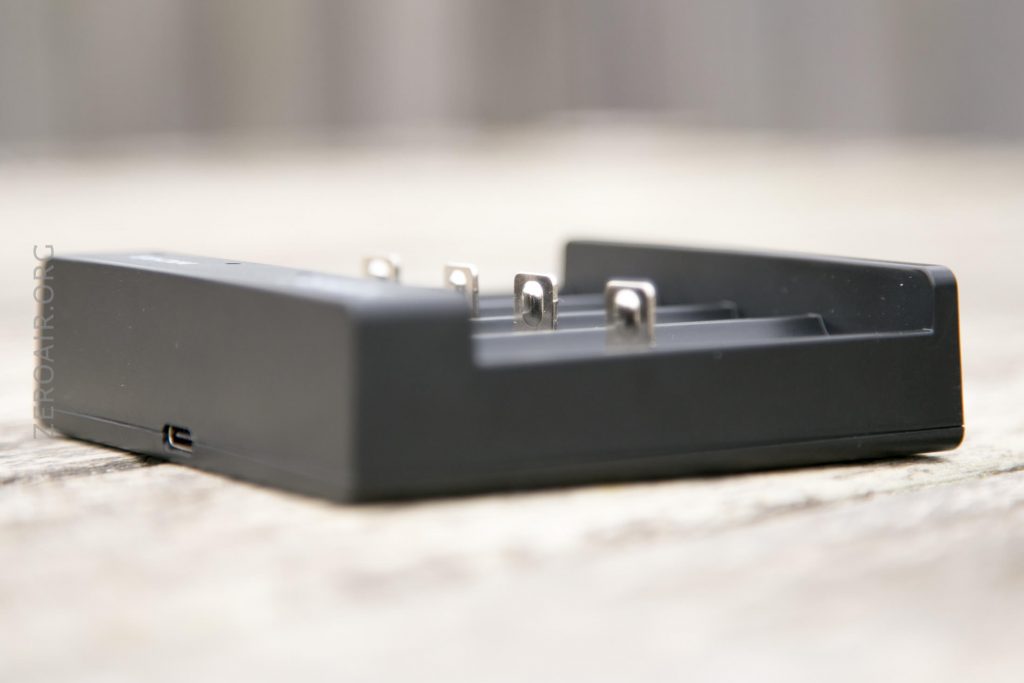 A close-up view of a black battery charger with four metal contacts, placed on a light-colored wooden surface. The side of the charger has a small port. The background is blurred.