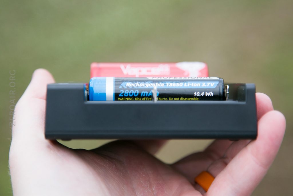 A hand holds a black battery charger with a blue and black rechargeable 18650 Li-ion battery labeled 2800 mAh and 3.7V. Another red battery is partly visible behind the charger.