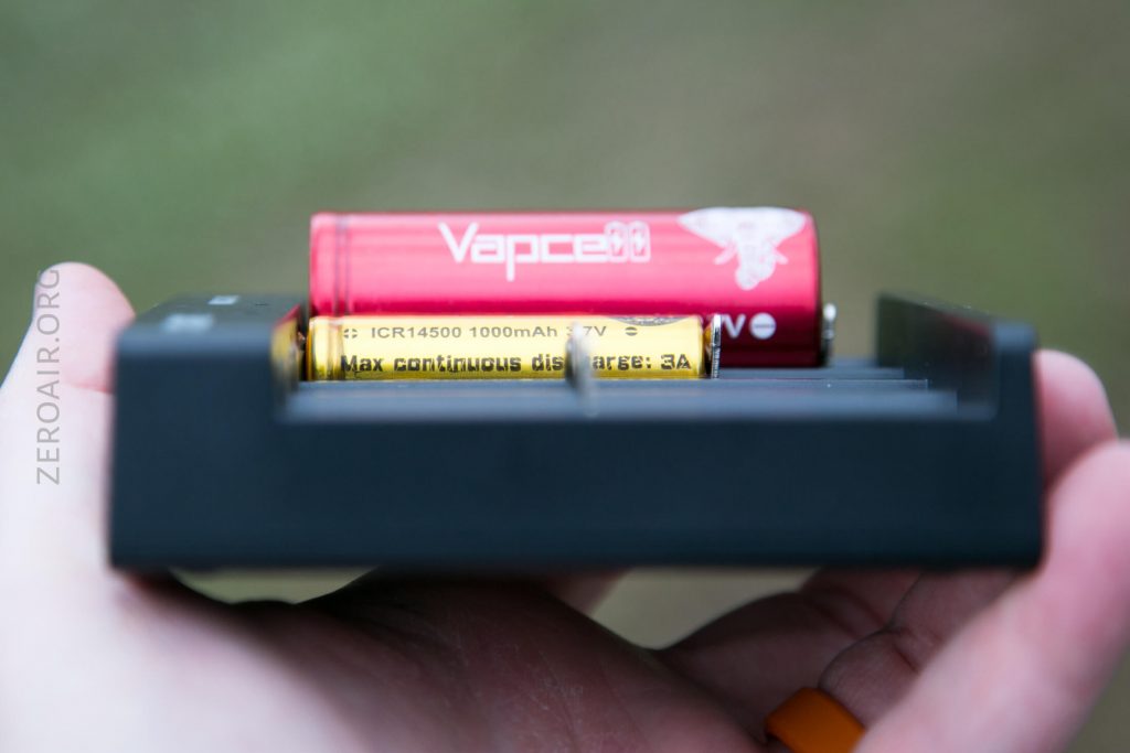 A close-up of a hand holding a battery charger with two cylindrical batteries, one yellow and one red, partially inserted. The charger is black and the background is blurred.