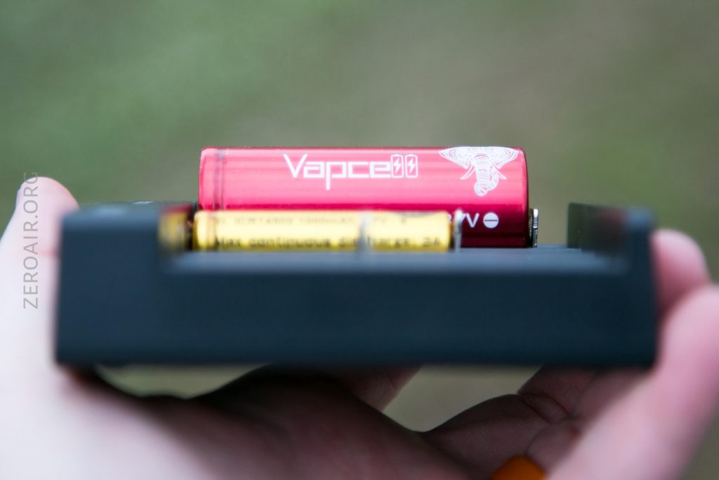 A close-up of a persons hand holding a black battery charger with a red Vapcell battery and a yellow battery placed inside it, against a blurred outdoor background.