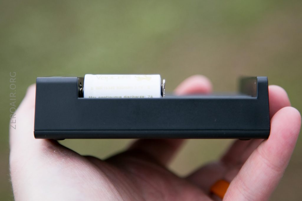 A close-up of a hand holding a black rectangular battery charger with a single cylindrical battery inserted. The battery has a partially visible label reading Vapcell. The background is blurred.