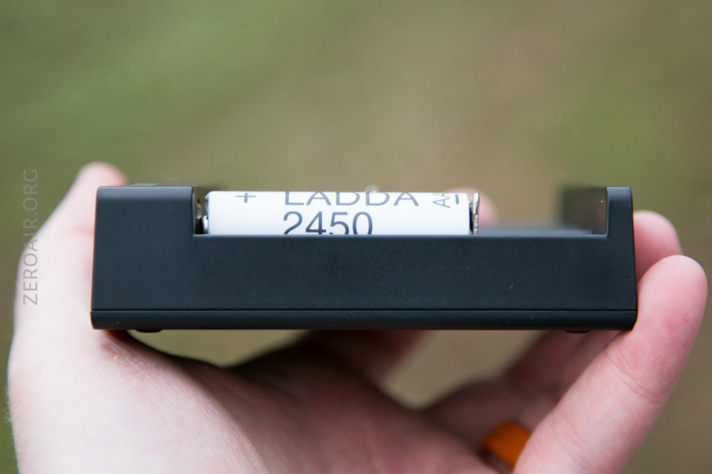 A hand holding a black battery charger with a white rechargeable battery labeled LADDA 2450 partially visible inside. The background is blurred.