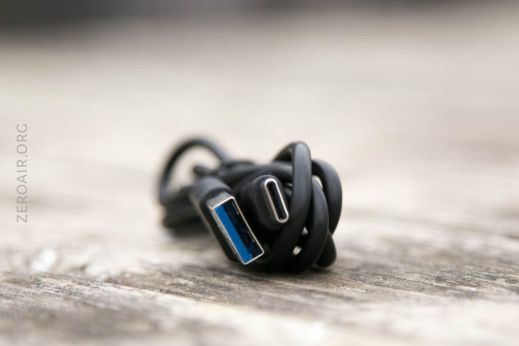 A black USB cable, featuring a USB-A connector on one end and a USB-C connector on the other, is coiled and resting on a wooden surface. The background is blurred, and ZEROAIR.ORG is visible on the left side.
