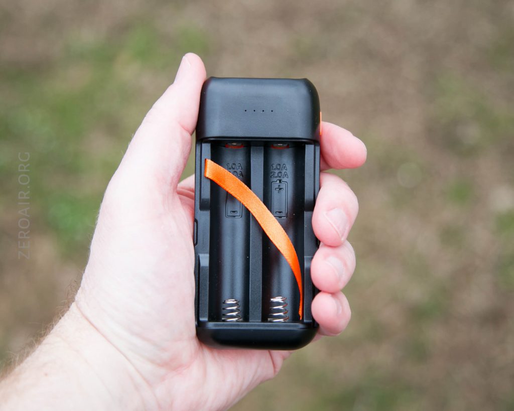 A hand holding a black battery charger with two empty slots and an orange ribbon inside. The background is out of focus, and the words ZEROAIR.ORG are faintly visible on the left side.