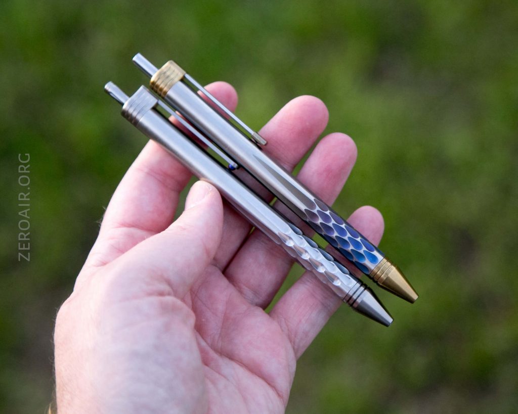 A hand holding two metal pens with textured, hexagonal patterns on the barrels; one pen has gold-colored accents, the other has silver and blue accents. The background is out-of-focus greenery.