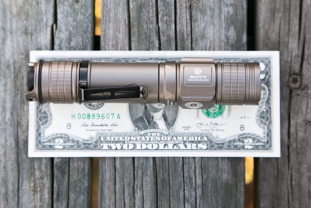 A bronze Brinyte brand flashlight is placed horizontally on top of a flat U.S. two-dollar bill, both resting on weathered wooden planks.