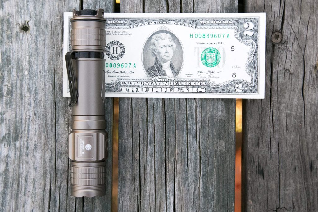 A metallic flashlight is placed next to a two-dollar bill on a weathered wooden surface, showing a size comparison between the flashlight and the banknote.
