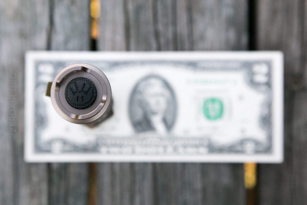 A close-up, top-down view of a metal object with a black circular end, placed vertically on a wooden surface, with a blurred US dollar bill underneath.