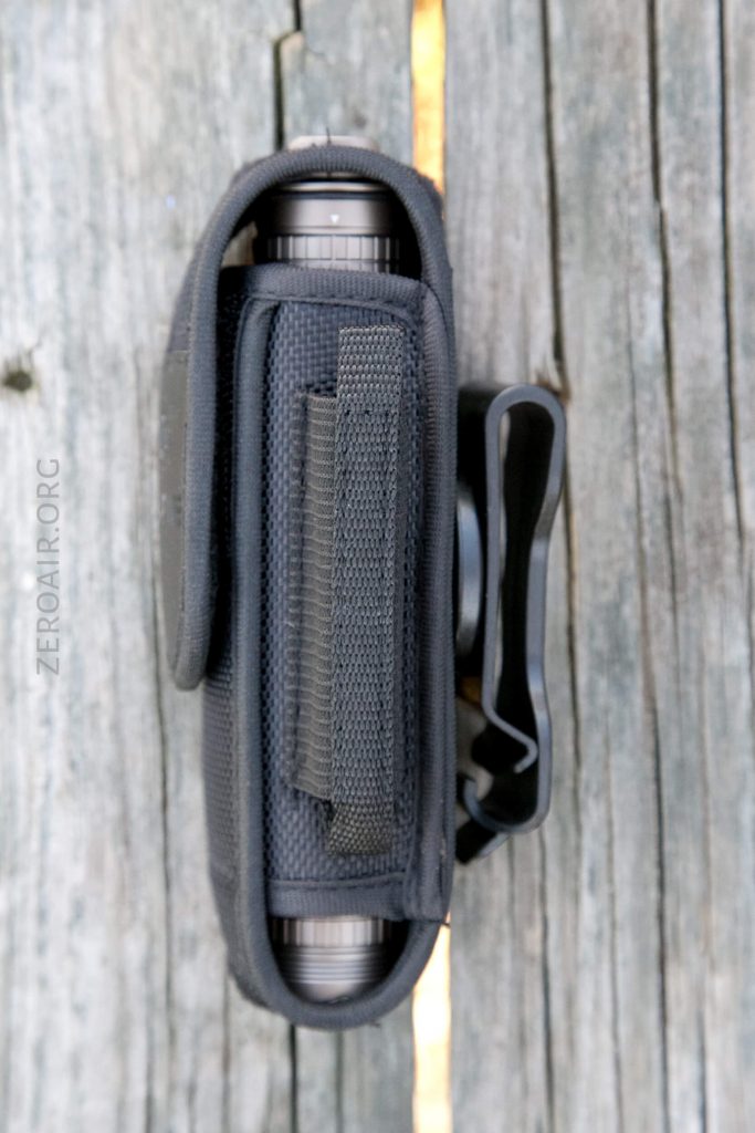 A black nylon pouch with a hook-and-loop strap and a metal belt clip holds a cylindrical object, photographed from above on a weathered wooden surface.