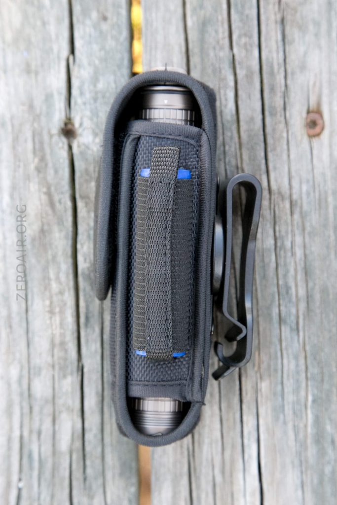 A black nylon case with a vertical strap and belt clip is holding a cylindrical object, placed on a weathered wooden surface.