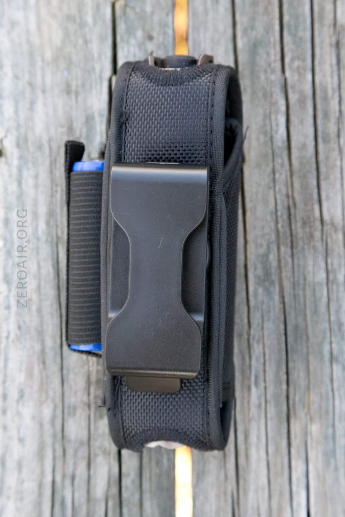 A black nylon pouch with a large metal clip attached, placed vertically on weathered wooden boards. A blue edge and some webbing detail are visible on the left side of the pouch.