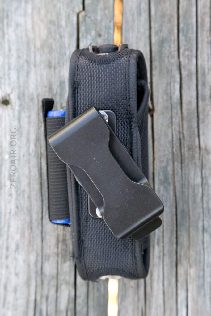 A black nylon holster with a metal belt clip is placed on a weathered wooden surface. The holster appears to be closed and has a visible elastic band on the side. The text ZEROAIR.ORG is visible on the left.
