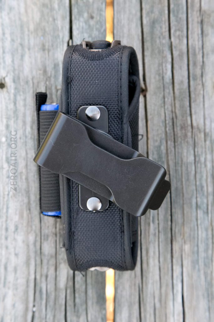 A black nylon pouch with a metal belt clip attached, positioned vertically on a weathered wooden surface. The pouch has visible stitching, snaps, and a blue accent on the side.