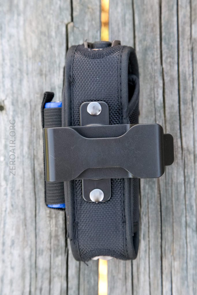 A black nylon pouch with a metal clip is secured to weathered wooden planks. The fabric has visible stitching and two metal rivets on the clip. Part of a blue object is partially visible inside the pouch.