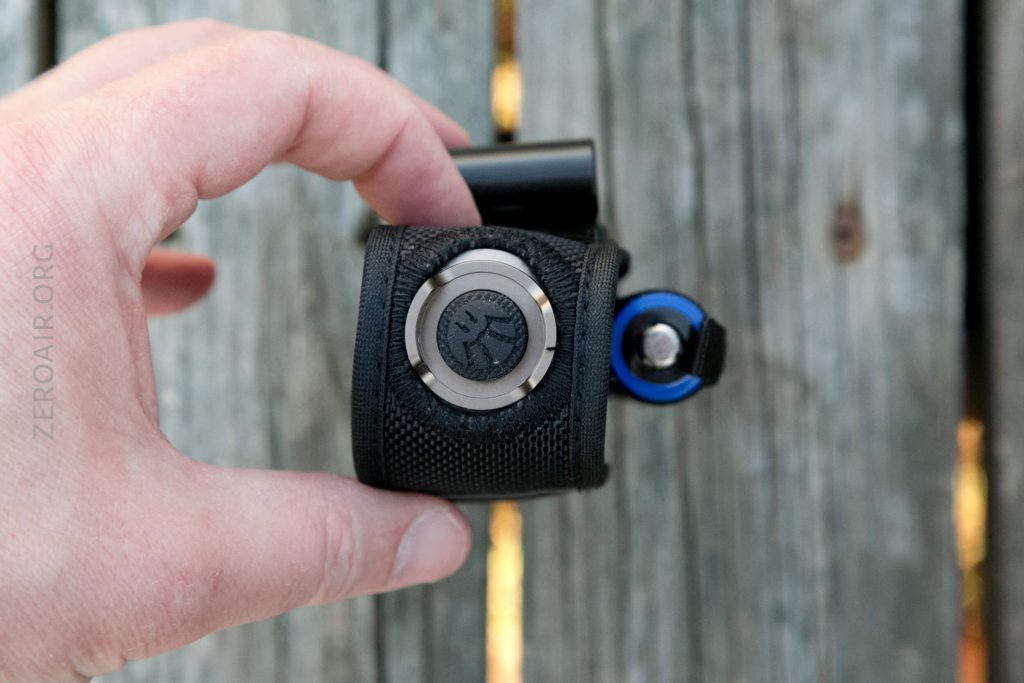 A hand holding a black fabric pouch with a circular silver and black button on top, resting on weathered wooden planks. A small blue object is partially visible attached to the pouch.