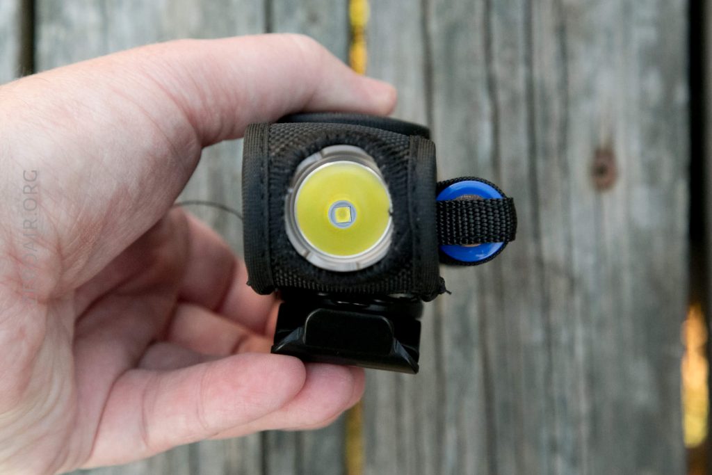 A hand holds a small black flashlight headlamp with a yellow LED at the center, a blue button on the side, and a Velcro strap, against a weathered wooden background.