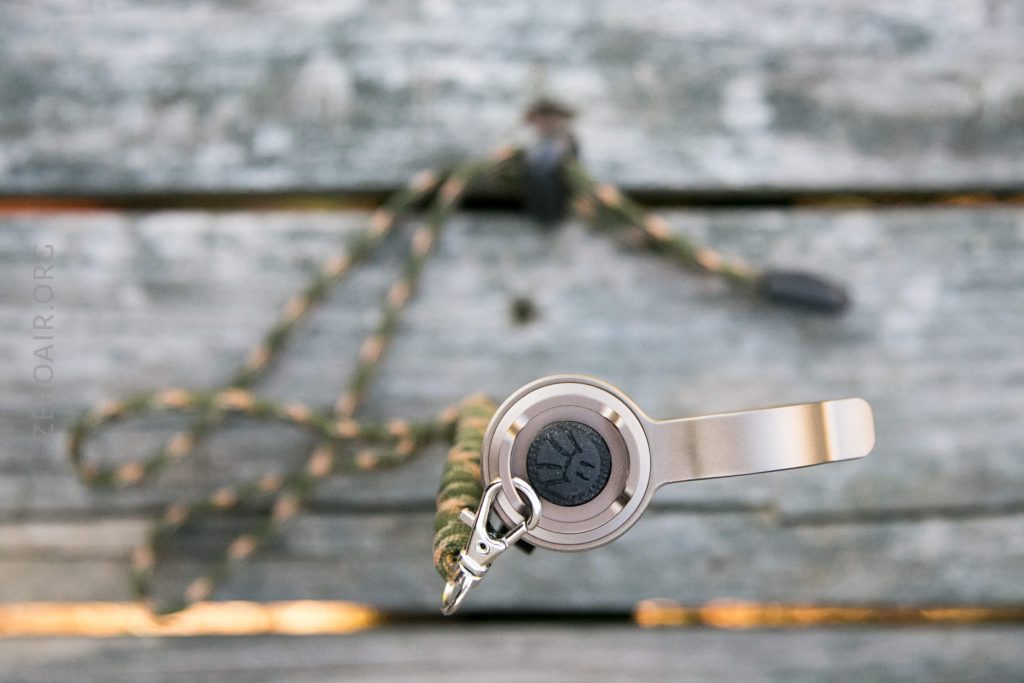 A close-up, top-down view of a metallic device attached to a green and brown braided lanyard, resting on weathered wooden boards. The device has a round black button or dial in the center.