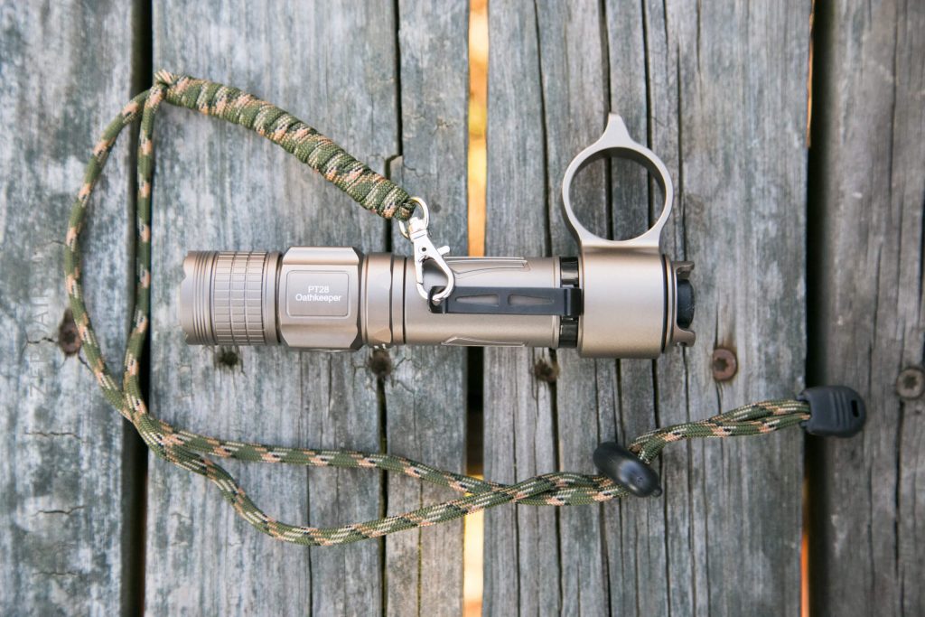 A metallic flashlight with a paracord lanyard attached is resting on weathered wooden boards. The flashlight has a ring near the end and engraved text on the side.