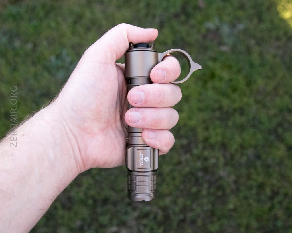 A person holding a metallic flashlight with a ring on the end, outdoors with grass in the background. The flashlight is gripped firmly in the right hand.