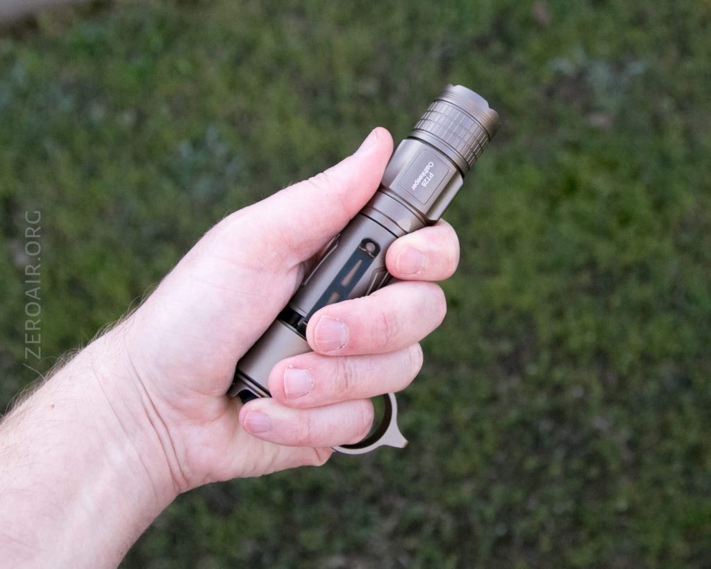 A left hand holds a metallic flashlight with a visible side switch and a knurled grip, against a grassy outdoor background. The flashlight features a trigger-like attachment near the thumb.