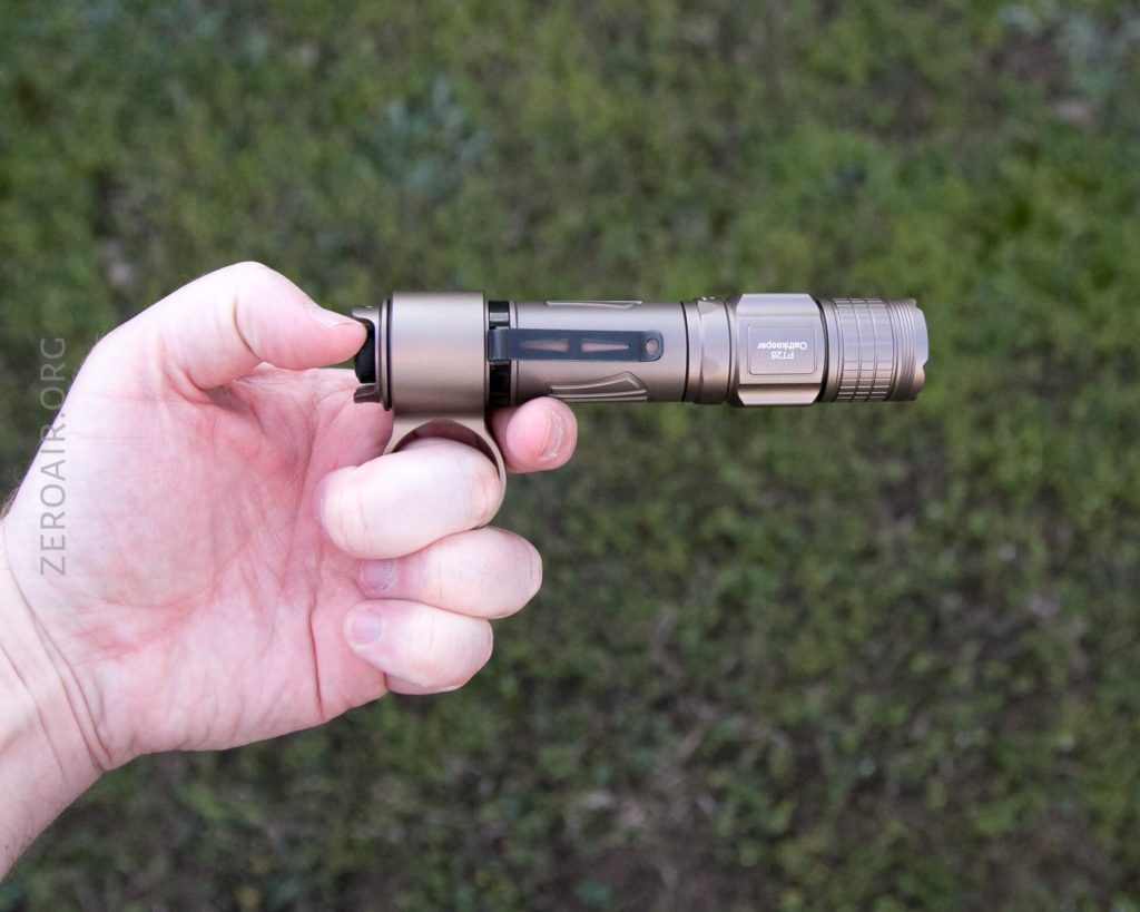 A hand holding a bronze-colored flashlight horizontally outdoors, with grass visible in the background. The flashlight has a textured grip and visible branding on the side.
