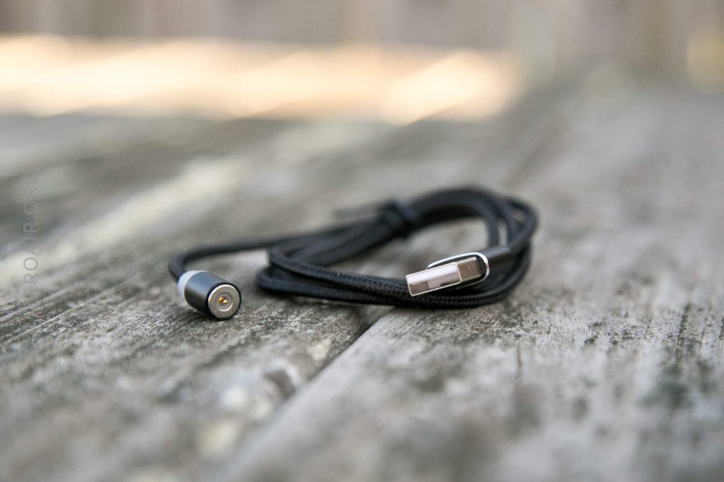 A black charging cable with a USB connector and a round magnetic power tip is coiled and resting on a weathered wooden surface.