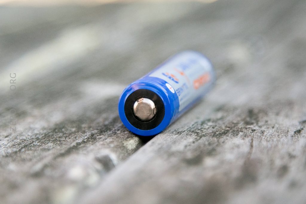 A close-up view of a single blue AA battery lying on its side on a weathered wooden surface. The positive terminal faces the camera, and the batterys label is partially visible.