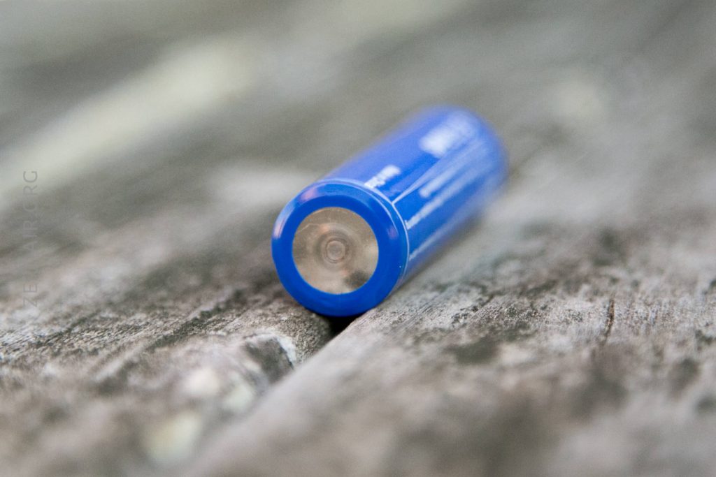 A close-up of a blue cylindrical battery lying on its side on a weathered wooden surface, with the focus on the terminal end of the battery.